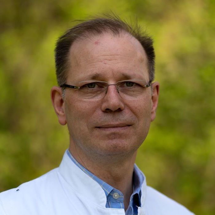 Ein Mann mittleren Alters mit Brille und weißem Kragen steht im Freien vor einem unscharfen grünen Hintergrund und blickt mit neutraler Miene in die Kamera. Er stellt das Nierenzentrum in Berlin Zehlendorf dar.