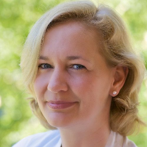 Eine Frau mit kurzen blonden Haaren und heller Haut, die Perlenohrringe und ein weißes Oberteil trägt, lächelt sanft in die Kamera. Das verschwommene Grün im Hintergrund suggeriert eine ruhige Umgebung in der Nähe des Nierenzentrums in Berlin Zehlendorf.