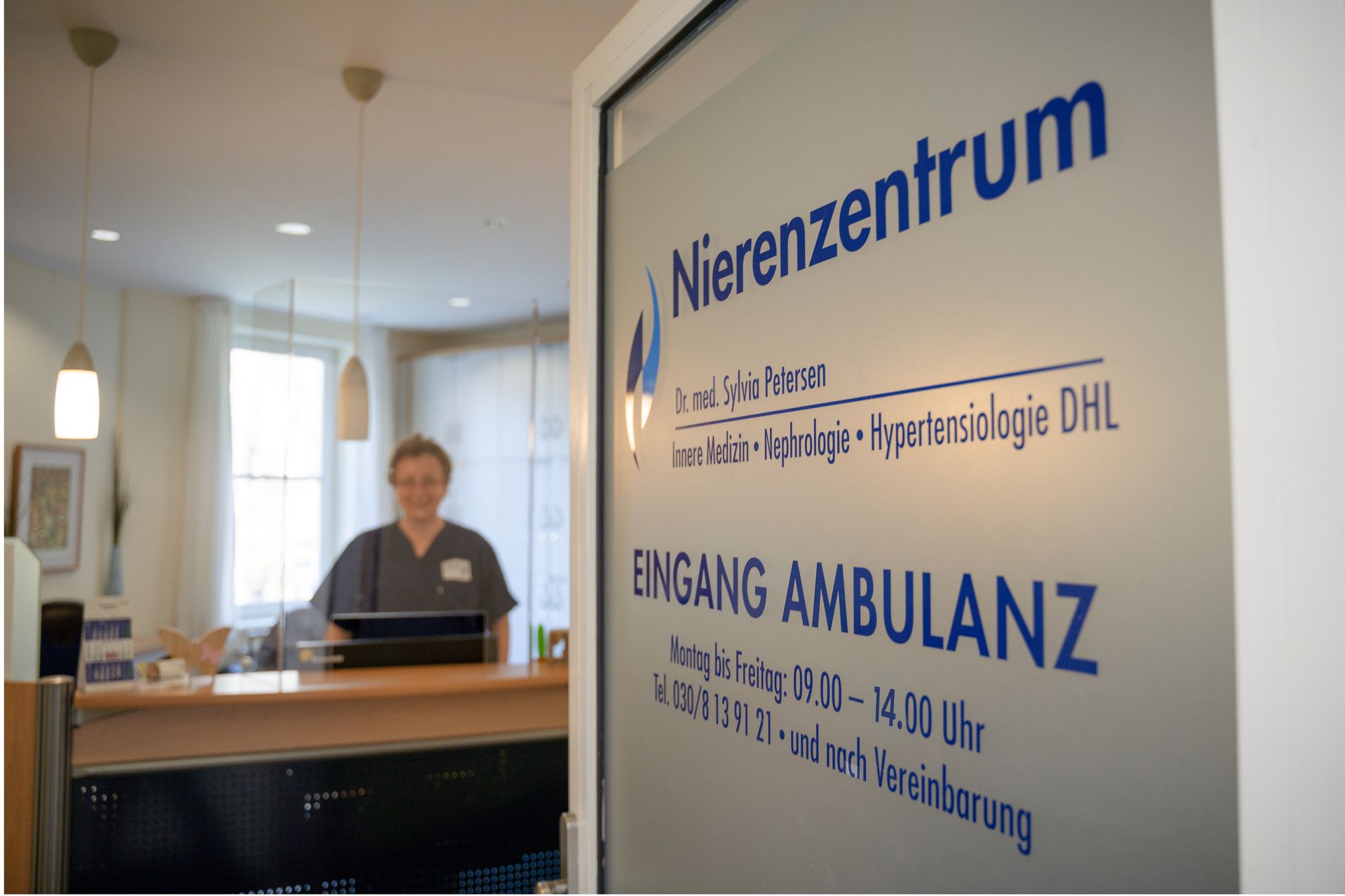 Empfangsbereich des Nierenzentrums Zehlendorf. Eine Mitarbeiterin steht hinter dem Schreibtisch im Hintergrund. Die Glastür im Vordergrund zeigt die Ambulanz und die Öffnungszeiten der nephrologischen Klinik in deutscher Sprache an.