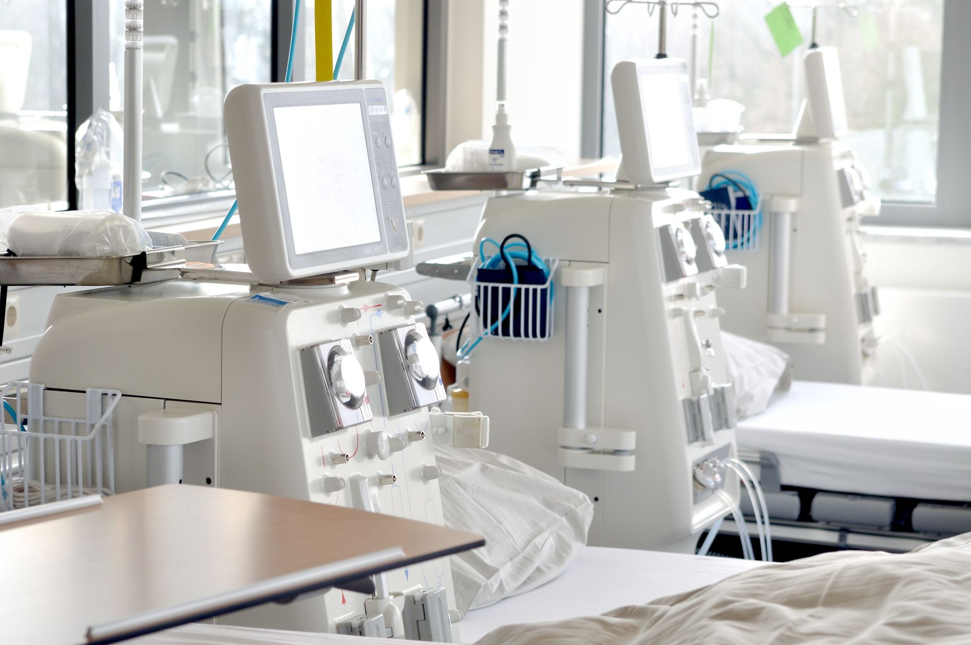 Eine Reihe von Dialysegeräten mit Monitoren und Schläuchen steht neben leeren Krankenhausbetten in der Feriendialyse Berlin Nierenzentrum Zehlendorf, Sonnenlicht strömt durch große Fenster in diese helle medizinische Einrichtung.