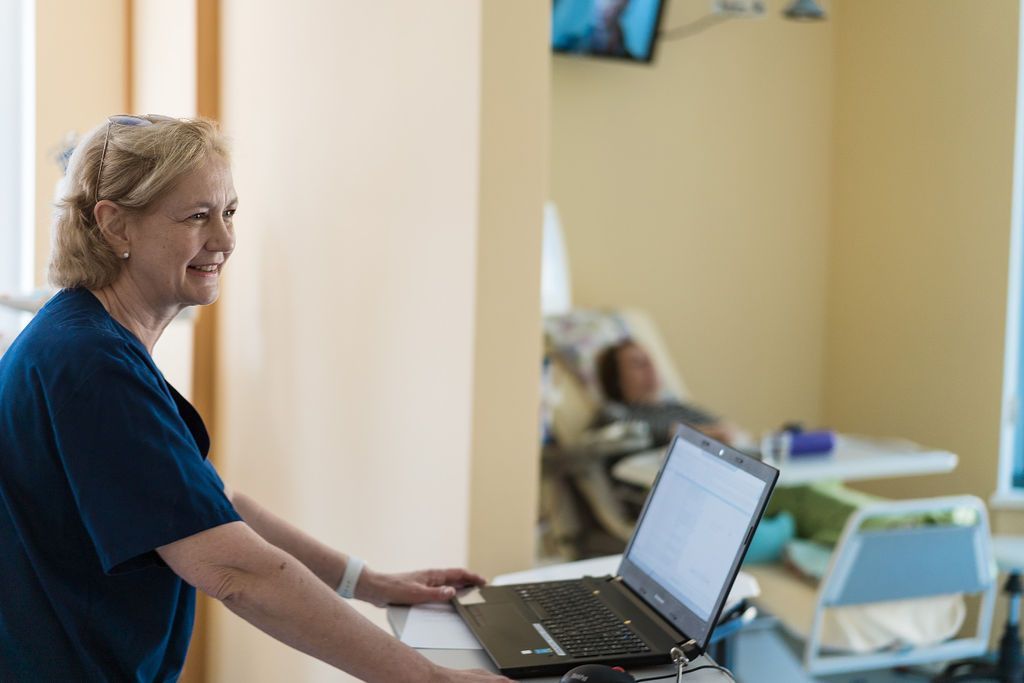 Eine Krankenschwester in blauem Kittel lächelt, während sie einen Laptop auf einem medizinischen Wagen in der Feriendialyse Berlin Nierenzentrum Zehlendorf benutzt; im Hintergrund ruht ein Patient bequem in einem hell erleuchteten Krankenhauszimmer.