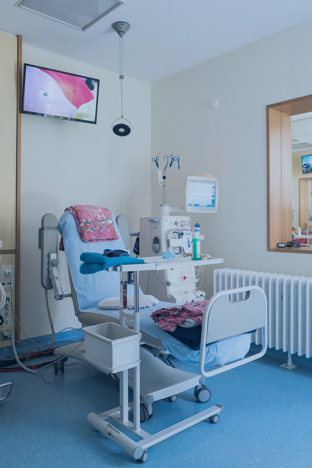 Ein modernes Krankenhauszimmer für die Hämodialyse im Nierenzentrum Zehlendorf verfügt über einen Dialysestuhl, medizinische Geräte, einen Monitor, gefaltete Wäsche, einen an der Wand montierten Fernseher und einen großen Spiegel, der einen Teil des Raumes reflektiert.