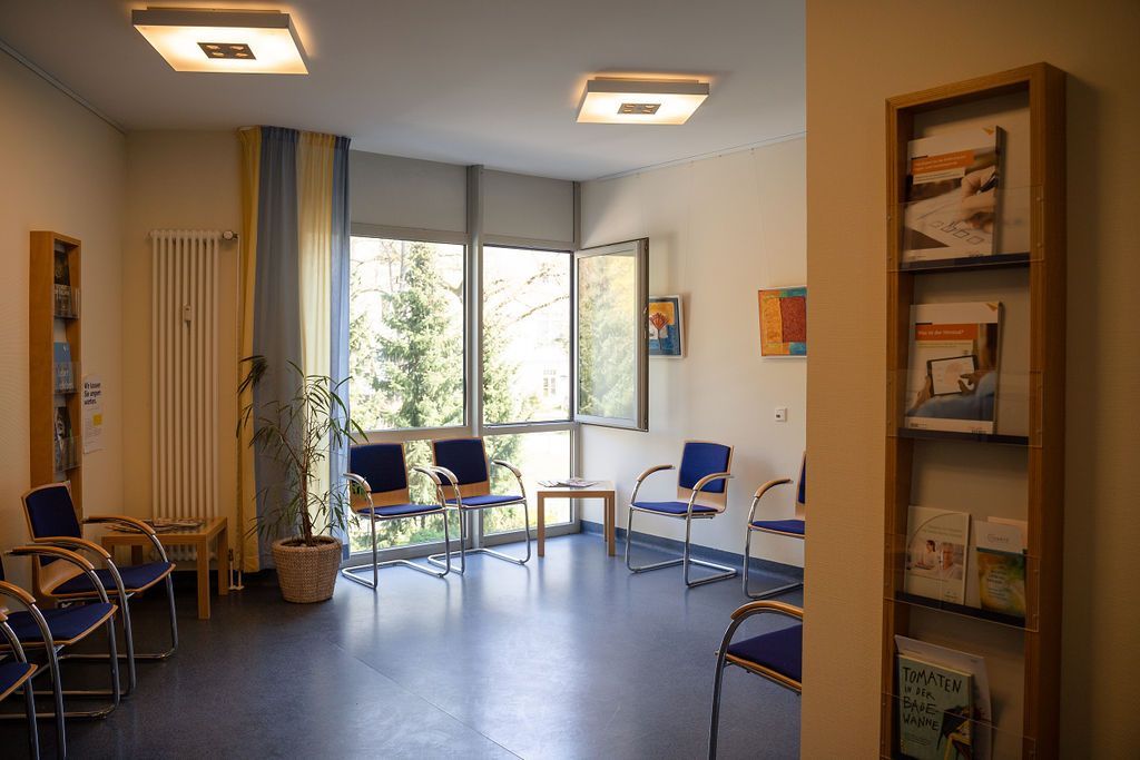 Im Wartezimmer des Nierenzentrums Zehlendorf stehen blaue Stühle in einem Kreis, eine Topfpflanze, große Fenster mit Tageslicht und ein Wandregal mit Broschüren. Der Raum der Ambulanz ist sauber und ruhig.