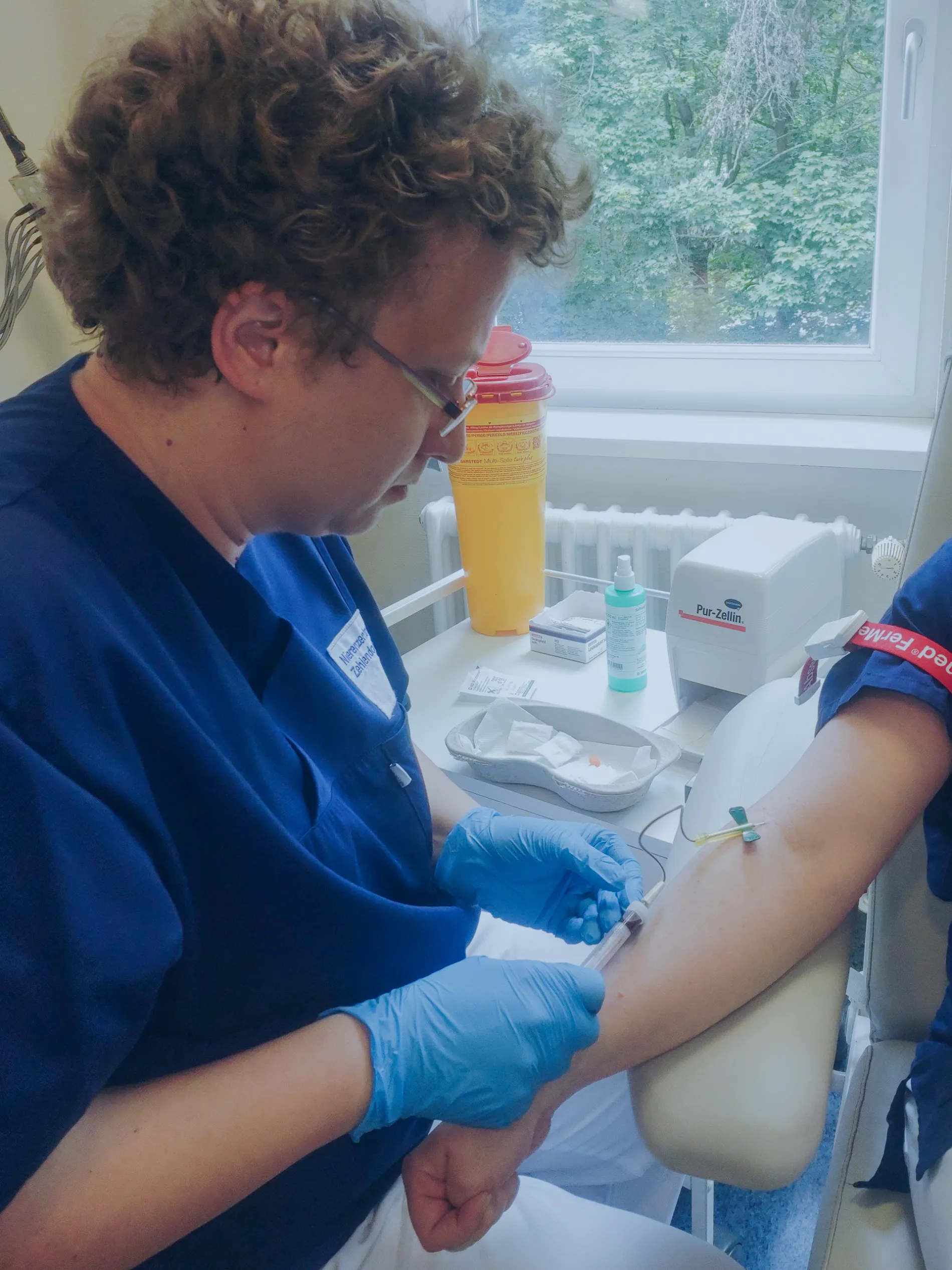 Eine medizinische Fachkraft in blauem Kittel und mit Handschuhen entnimmt einem Patienten mit einer Spritze Blut aus dem Arm in einem medizinischen Büro in der Hämodialyse im Nierenzentrum Zehlendorf, während im Hintergrund Material auf einem Tisch liegt und ein Fenster zu sehen ist.